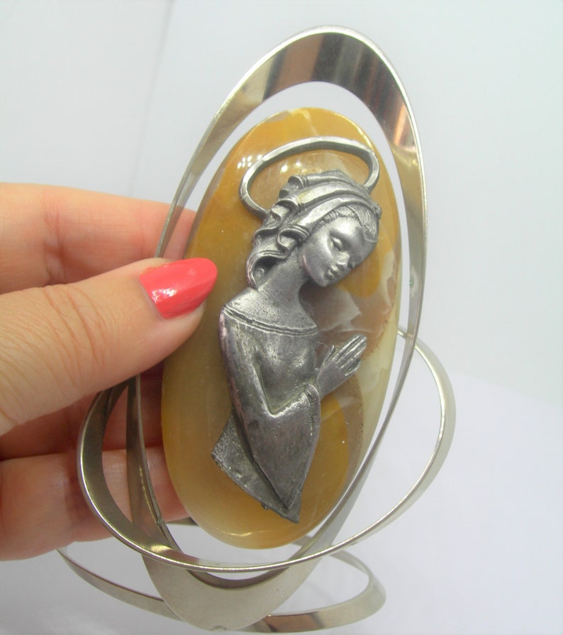 May include: A silver oval frame with a yellow oval stone inside. The stone has a silver figure of a woman with her hands clasped in prayer.