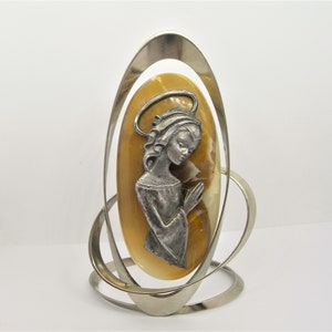 May include: A silver metal stand with a circular design holds a yellow oval cameo with a silver image of a woman with a halo praying.