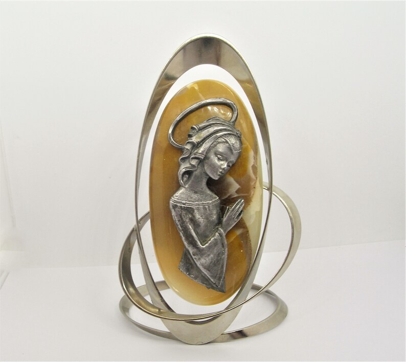 May include: A silver metal stand with a circular design holds a yellow oval cameo with a silver image of a woman with a halo praying.