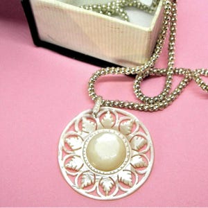 May include: A silver-toned necklace with a large, round pendant featuring a central pearl-like bead and a decorative leaf design. The necklace chain is a beaded style, and the pendant is resting on a pink surface.
