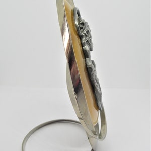 May include: A silver oval-shaped frame with a beige background and a pewter image of a woman's face. The frame has a silver stand.