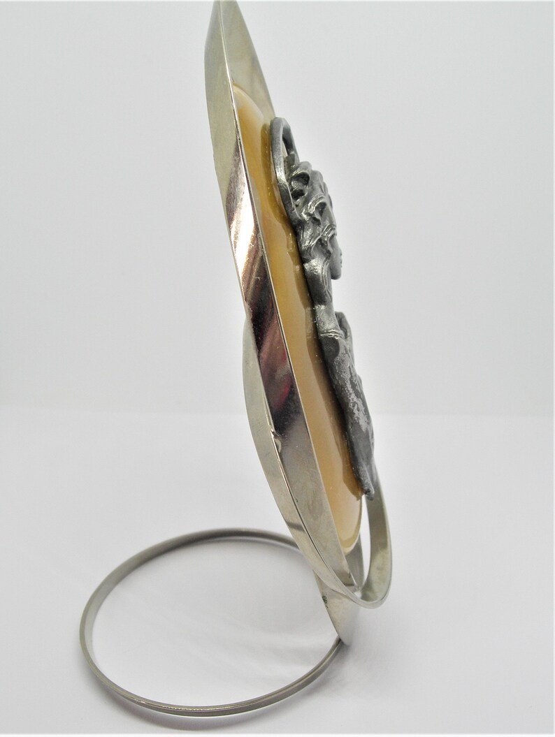 May include: A silver oval-shaped frame with a beige background and a pewter image of a woman's face. The frame has a silver stand.