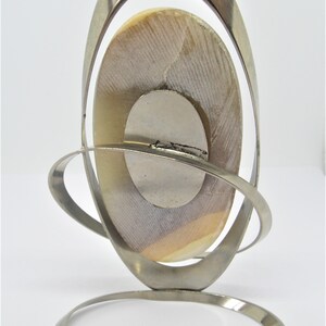 May include: A silver metal stand with a circular base and a curved, oval-shaped frame. A round, white and brown shell is displayed in the frame.