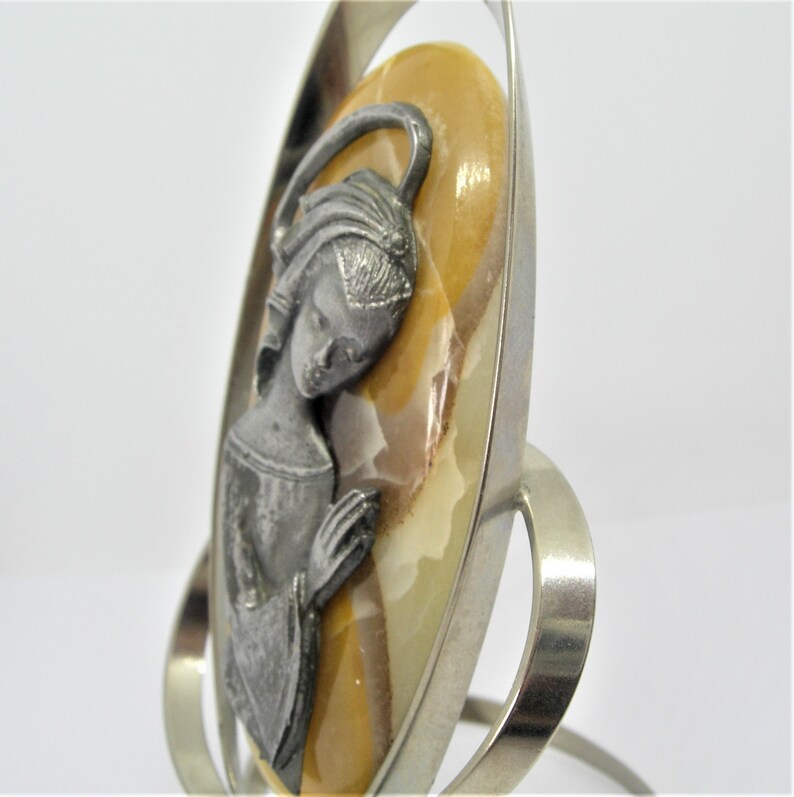 May include: A silver metal stand with a round, oval-shaped frame holding a religious icon. The icon is a relief of a woman with a halo, made of pewter and set against a yellow and brown agate stone.