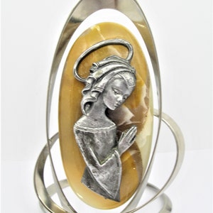 May include: A silver metal stand with a swirling design holds a light brown oval-shaped stone with a silver figurine of a woman praying on it.