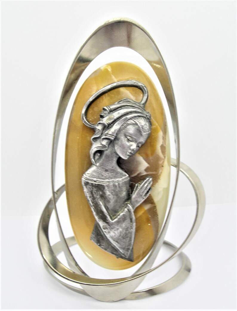 May include: A silver metal stand with a swirling design holds a light brown oval-shaped stone with a silver figurine of a woman praying on it.