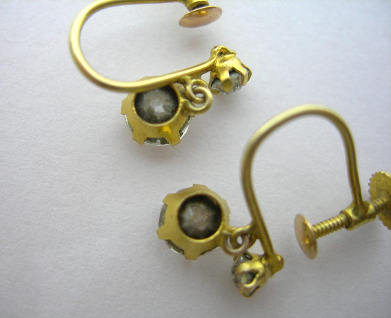 May include: A pair of gold earrings with a small clear stone on each earring. The earrings have a screw-back fastening.