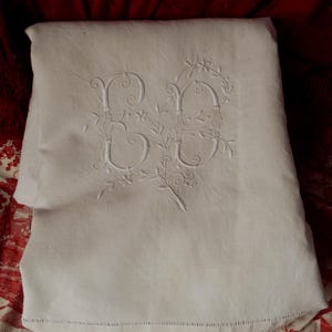 May include: A folded white linen textile featuring intricate white-on-white embroidery of the letters "ED" intertwined with delicate floral and vine motifs. The fabric displays a hemstitched border along the bottom edge, showcasing fine needlework.