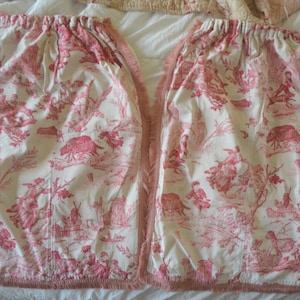 May include: Two matching fabric panels featuring a red toile de jouy pattern on a cream background. The design depicts pastoral scenes with figures, animals, and structures. Each panel has a gathered top edge and a frayed, raw-edge trim along the side.