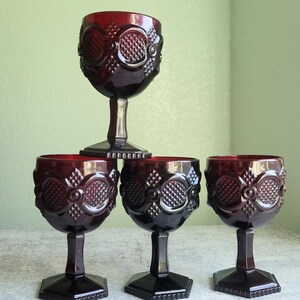 Può includere: Cinque calici da vino rosso rubino. Ogni calice presenta un design ornato e strutturato sulla coppa, con stelo e base neri. I calici sono disposti su una superficie chiara, davanti a un muro verde chiaro. Altezza: circa 15 cm.
