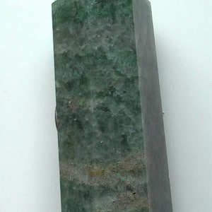 Puede incluir: Una punta de cristal verde con una parte superior puntiaguda y una base plana. El cristal tiene una apariencia nublada y transl&uacute;cida con un tono verde.