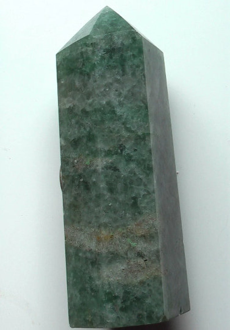 Puede incluir: Una punta de cristal verde con una parte superior puntiaguda y una base plana. El cristal tiene una apariencia nublada y transl&uacute;cida con un tono verde.