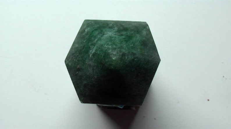 Puede incluir: Un cristal hexagonal facetado de color verde oscuro. El cristal tiene una textura rugosa y natural.
