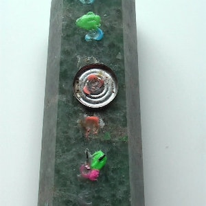 Puede incluir: Un obelisco de piedra verde con varios adornos coloridos, incluyendo un disco met&aacute;lico redondo con un centro rojo.