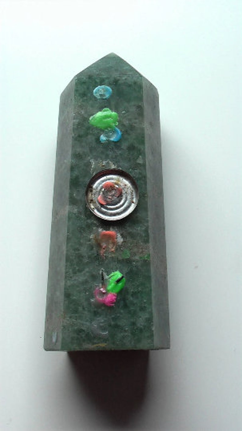 Puede incluir: Un obelisco de piedra verde con varios adornos coloridos, incluyendo un disco met&aacute;lico redondo con un centro rojo.