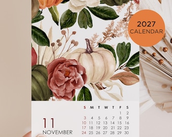 May include: A 2027 calendar page for November featuring a watercolor illustration of pumpkins, dark green leaves, and a large red flower. The calendar grid displays the days of the month with the text 11 NOVEMBER printed in black and red ink.