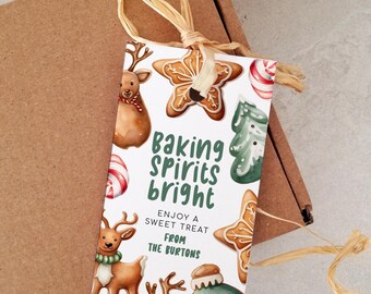 May include: A rectangular gift tag with the text "Baking Spirits Bright" and festive illustrations of reindeer, gingerbread cookies, and candy canes. The tag is tied with raffia and rests on a brown cardboard box.