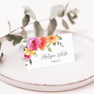 May include: White place card with watercolor floral design featuring pink, orange, and yellow flowers. The text on the card reads "Adelyne White, Table 1".