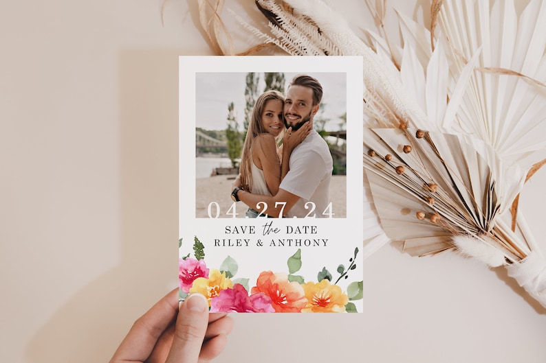May include: Save the date card with a floral design featuring pink, yellow, and orange flowers. The card has a photo of a couple and the text "04.27.24 Save the Date Riley & Anthony".