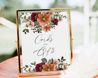 May include: A rectangular gold-toned metal frame holding a white sign with the black text Cards AND Gifts. The sign is decorated with watercolor-style floral arrangements at the top and bottom featuring burgundy, burnt orange, and green foliage.