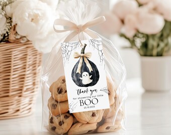May include: A clear cellophane bag tied with a beige ribbon, filled with chocolate chip cookies. A tag with a black pumpkin, ghost, and spiderwebs reads "Thank you for showering our little BOO 25.10.2025". The background features white flowers in a basket.