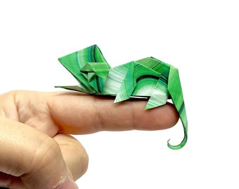Może przedstawiać: Szczegółowy słoń origami, wykonany z zielonego papieru we wzory, spoczywa na palcu. Papierowy słoń ma zakręcony trąbę i skomplikowane fałdy, demonstrując sztukę składania papieru. Tło jest jednolicie białe.