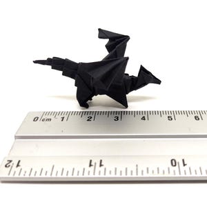 Può includere: Una figura di drago in origami nero posizionata sopra un righello di metallo. Il righello mostra misure sia metriche che imperiali, con il drago che misura circa 6,5 cm di lunghezza dalla testa alla punta della coda appuntita.
