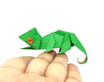 Może przedstawiać: Zielony kameleon origami z pomarańczowymi akcentami, spoczywający na dłoni. Papierowa rzeźba ma szczegółowe fałdy, ukazujące kształt jaszczurki. Oko kameleona to mały pomarańczowy kwadrat. Tło jest jednolicie białe.