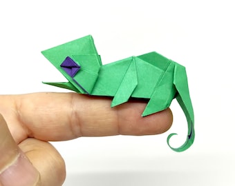 Może przedstawiać: Zielony origami kameleon z fioletowym okiem, siedzący na palcu.