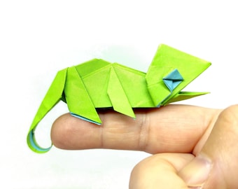 Może przedstawiać: Jasnozielony kameleon origami ze zwiniętym ogonem i niebieskimi akcentami. Papierowa rzeźba spoczywa na palcu, prezentując szczegółowe fałdy i geometryczne kształty rzemiosła. Okiem kameleona jest mały niebieski romb.