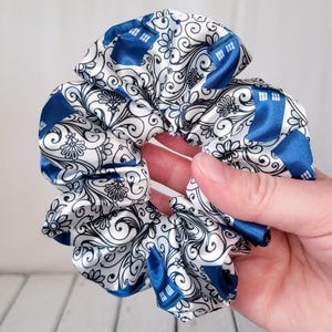 May include: A white and blue patterned scrunchie with a blue TARDIS design. The scrunchie is made of satin fabric and has a ruffled texture.