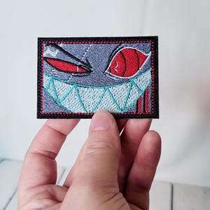 May include: Embroidered patch featuring a cartoon demon face with red eyes and a toothy grin. The patch is outlined in black and has a red and white border.
