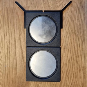 May include: Two black square frames with a silver circular disc in each frame. The frames are attached to each other and have a small black tab on each side.