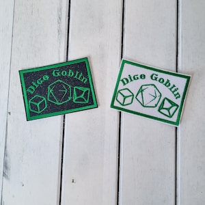 May include: Two embroidered patches with a green border and the text "Dice Goblin" in green. The left patch has a black background and green dice. The right patch has a white background and white dice.