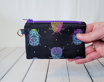 May include: A small black zippered pouch with a rainbow beetle pattern. The pouch has a purple zipper and a gold ring.
