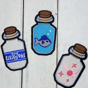 May include: Three embroidered patches shaped like glass bottles with cork tops. The first bottle is blue and contains a blue fish with white eyes. The second bottle is white and has a blue label with the text "Milk" on it. The third bottle is white and contains a pink fairy with pink wings.