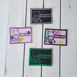 May include: Four embroidered patches featuring a cartoon cat holding a dice with the text "Dungeon Meowster" in black and white with a glittery background. The patches are in different colours: black, purple, pink, and green.