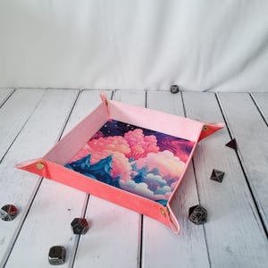 May include: A pink felt dice tray with a colorful fantasy landscape print. The tray has gold metal corners and is designed to hold dice.