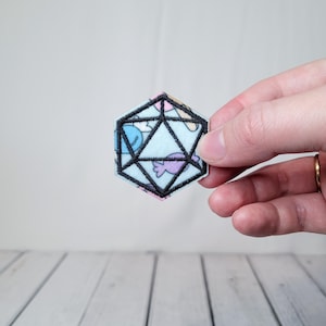 May include: A blue and white fabric patch with a black outline of a twenty-sided die.