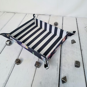 May include: A black and white striped dice rolling tray with a floral pattern on the outside. The tray is made of fabric and has a square shape. There are several dice scattered around the tray.