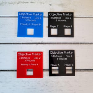 Puede incluir: Cuatro marcadores de objetivo para un juego de mesa. Dos son negros, uno es azul y uno es rojo. Cada marcador tiene el texto "Marcador de objetivo" y detalles sobre defensa, tamaño y heridas. Los marcadores también indican a qué jugador son amistosos.