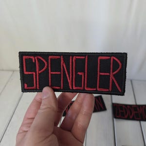May include: A rectangular embroidered patch with a black background and the word SPENGLER stitched in red capital letters. The patch features a thick black border and a textured fabric surface, with other similar patches visible in the background.