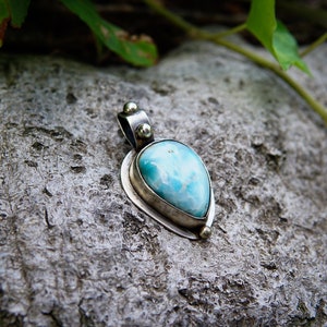 May include: A teardrop-shaped pendant with a blue and white gemstone set in silver. The pendant is on a rough, gray rock.