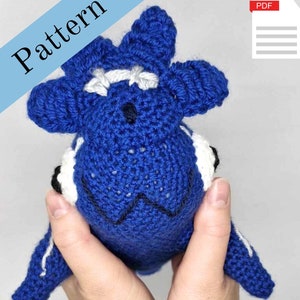 May include: A crocheted blue whale amigurumi toy with white and black details. The whale has a friendly face with black eyes and a white mouth. The toy is being held by a person's hands.
