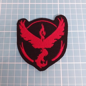 May include: Red and black embroidered patch featuring the Pokemon logo, a stylized bird with wings outstretched, inside a circle with a flame design.