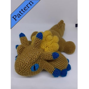 May include: A crocheted plush toy in the shape of a dragon. The dragon is brown with yellow accents and blue eyes. The dragon is lying on its side with its head resting on the ground.