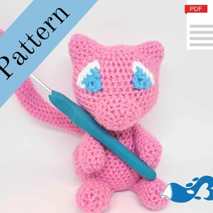以下が含まれることがあります： 青い目をした白い尻尾を持つポケモンのキャラクターに似ているピンクの毛糸で編まれたアミグルミのおもちゃ。おもちゃはかぎ針で持たれています。画像には「パターン」と「PDF」のテキストが含まれています。