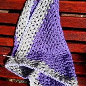 May include: A crocheted baby blanket with a purple and gray color scheme. The blanket is made with a granny square pattern and has a white border.