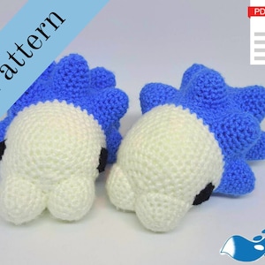 Puede incluir: Dos juguetes amigurumi de crochet azul y blanco. Los juguetes tienen forma de criatura marina con una espalda espinosa y un cuerpo redondo. Los juguetes están hechos con hilo blanco e hilo azul. Los juguetes están sobre una superficie blanca.