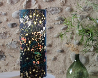 Puede incluir: Pantalla de lámpara cilíndrica con fondo azul oscuro y estampado floral en tonos blancos, rosas, naranjas y verdes. La pantalla está sobre una superficie blanca, con una pared de piedra al fondo.